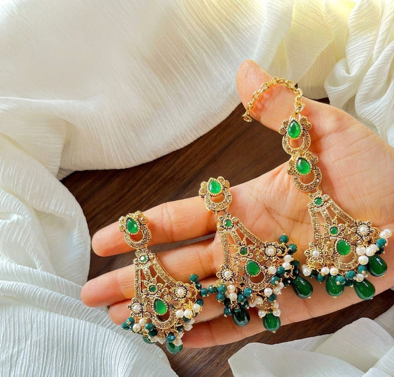 Hand-held display of antique gold chandbali earrings and maang tikka set with vibrant emerald green stones, white seed pearls, and dark green teardrop beads over white crushed fabric.Rich green focal stones contrasted against warm antique gold filigree and brilliant clear crystal trims.Delicate white seed pearls combined with teardrop emerald beads for an opulent dangling border.