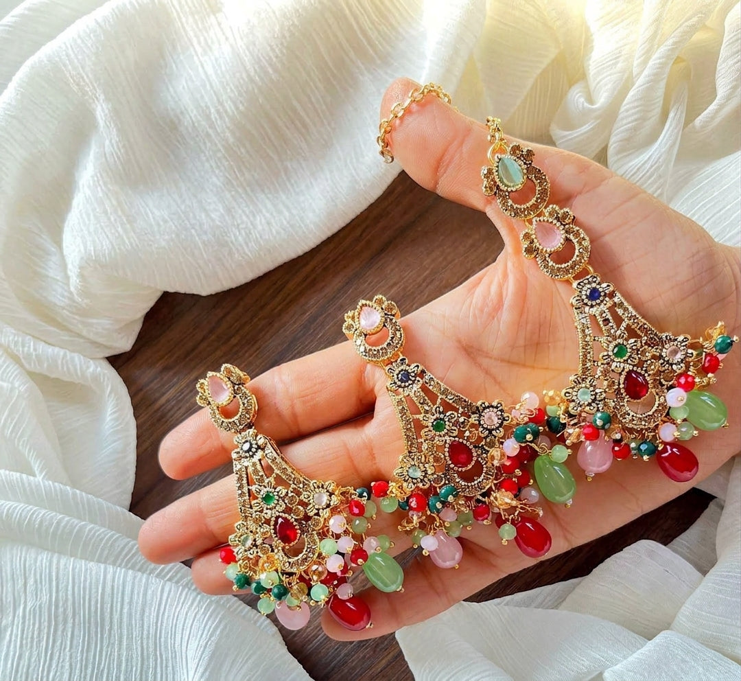 Hand-held display of antique gold chandbali earrings and maang tikka set with multi-colored stones, jade beads, and red teardrop tassels against white textured fabric.Intricate openwork motif accented with colorful center teardrop stones.Multi-layered hanging beads combining jade green, blush pink, and ruby red for dramatic movement.