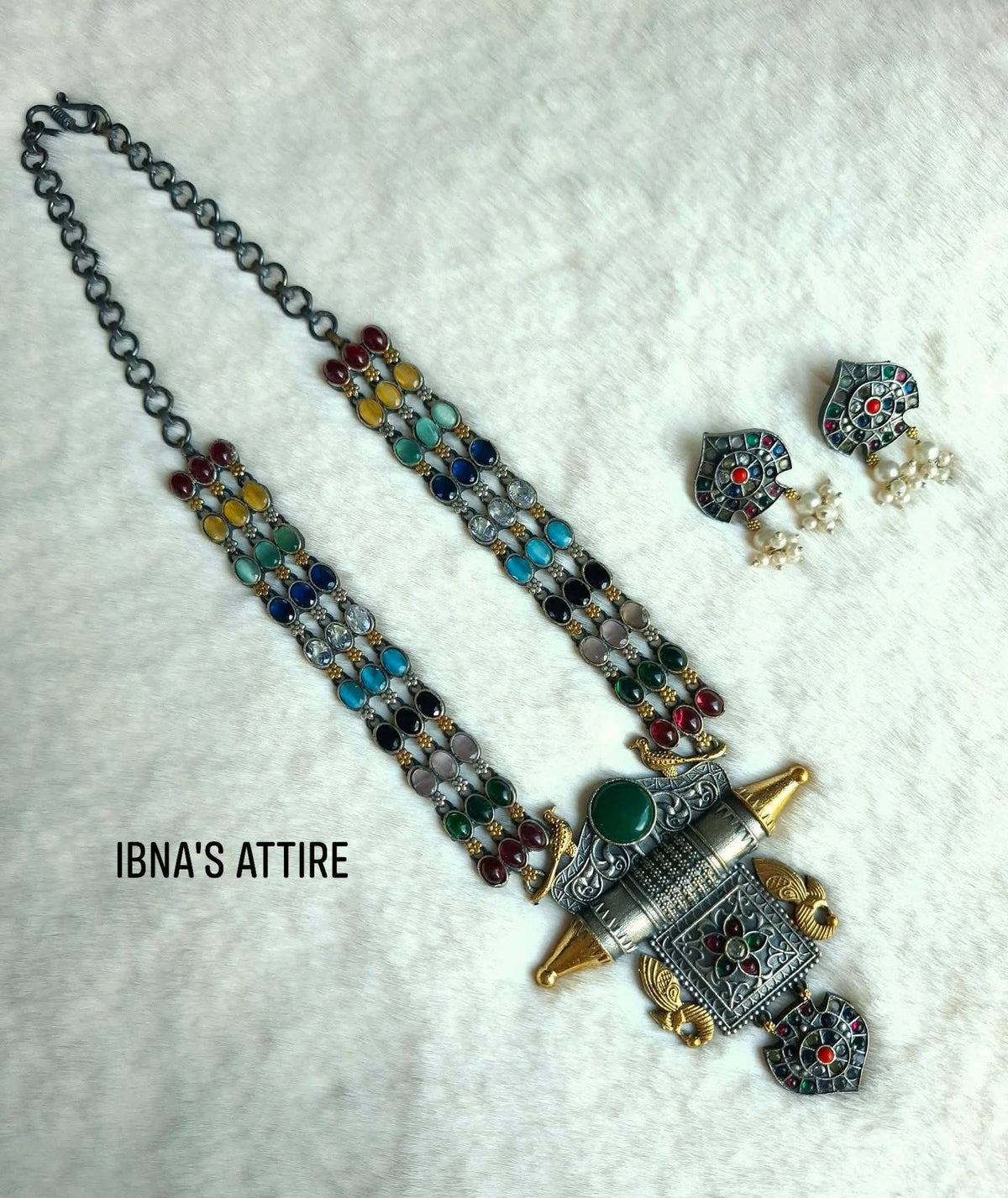 Display of a long oxidized German silver necklace with three multi-colored stone strands, dual-tone cylindrical taviz pendant, and matching heart-shaped pearl drop earrings on white plush background. Heart/shield-shaped stud earrings topped with colorful stone inlay and delicate pearl seed drops.Heavy cylindrical central motif featuring antique filigree engraving, bird motifs, and multi-stone inlay.Three parallel rows of bezel-set multi-color stones strung together for rich visual impact.  