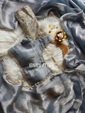Flat lay shot of a metallic slate-grey glass tissue silk saree draped over plush white faux fur, alongside a matching readymade tissue blouse with embroidered sheer net sleeves and scalloped gold lace borders.Light-reflecting glass tissue silk that shifts beautifully between silver-grey and muted slate blue shades.Intricate golden lace trim lined with fine pearl beads defining the full saree border and blouse frame.
