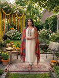 A woman in a lush garden courtyard wearing a cream-colored kurta with colorful geometric embroidery and a vibrant multicolor patterned shawl draped over her shoulders.