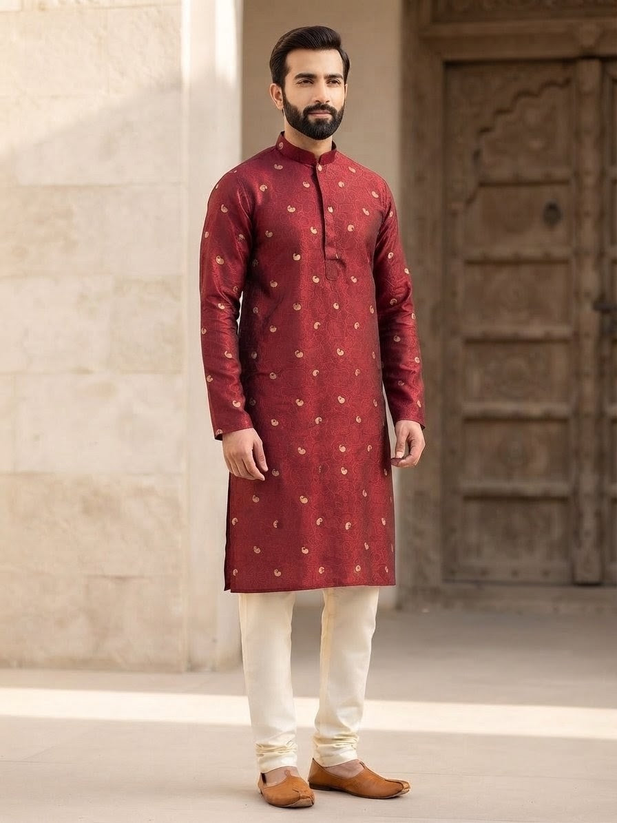 A man wearing a maroon long-sleeve kurta with small gold motifs, paired with off-white trousers and brown shoes, standing in front of a beige stone wall and wooden door.