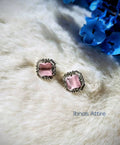 Silver-tone stud earrings with a square pink center stone and an outer American diamond frame, laid flat on a textured white fabric background near blue floral decor.