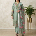 Model standing in a sage green embroidered lawn kameez with matching trousers and a printed floral dupatta next to a golden pot plant.Beautiful pastel embroidery that enhances the soft sage green background.