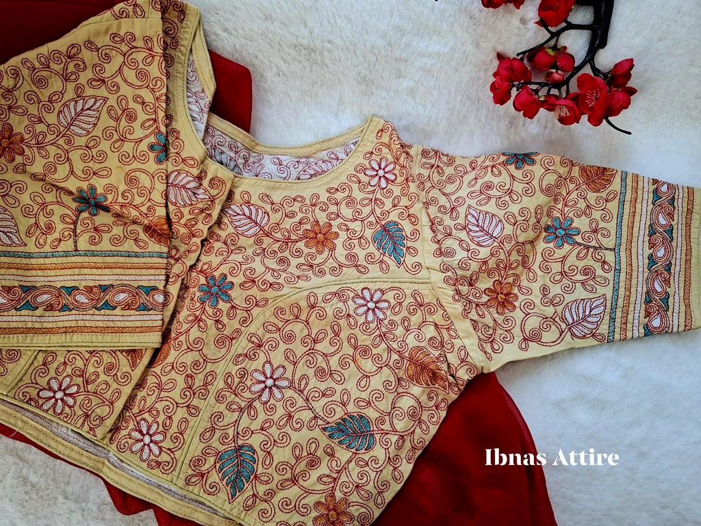 Beige multi color malai cotton blouse displayed flat over a deep red fabric on a soft white fur surface, highlighting fine Kantha stitch embroidery and a front hook closure.