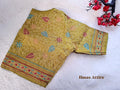 Olive green malai cotton blouse lying flat on a soft white faux-fur background, displaying detailed Kantha floral embroidery and a front hook closure.Round neckline with elbow-length/short sleeves bordered with neat stitching.Detailed running-stitch embroidery outlining floral trails and colorful leaves in red, blue, and gold tones.