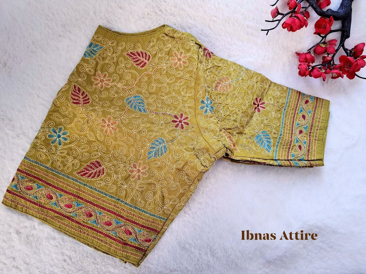 Olive green malai cotton blouse lying flat on a soft white faux-fur background, displaying detailed Kantha floral embroidery and a front hook closure.Round neckline with elbow-length/short sleeves bordered with neat stitching.Detailed running-stitch embroidery outlining floral trails and colorful leaves in red, blue, and gold tones.