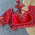 Bright red silk readymade blouse featuring real mirror embellishments, colorful Gamthi embroidery, Mumtaz neckline, 12-inch sleeves, and latkan ties displayed on a jute rug
