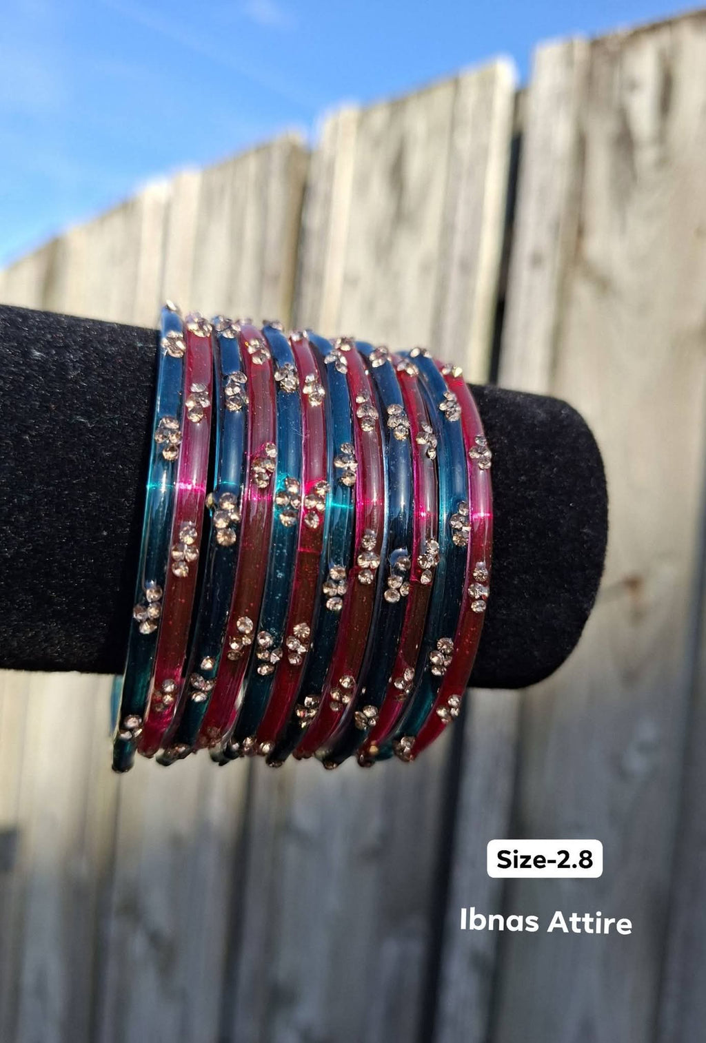Stack of translucent peacock teal and crimson red glass bangles with gold sequin accents displayed on a black velvet bar outdoors against a wooden fence.Alternating arrangement of dark teal and crimson magenta glass rings creating depth on the wrist.