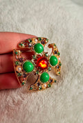 classic architectural cross structure detailed with filigree scrollwork ,Clusters of smaller multi-colored rhinestones and pearls add texture and depth,Bold green cabochons balanced by a vibrant central red stone brooch held by a hand