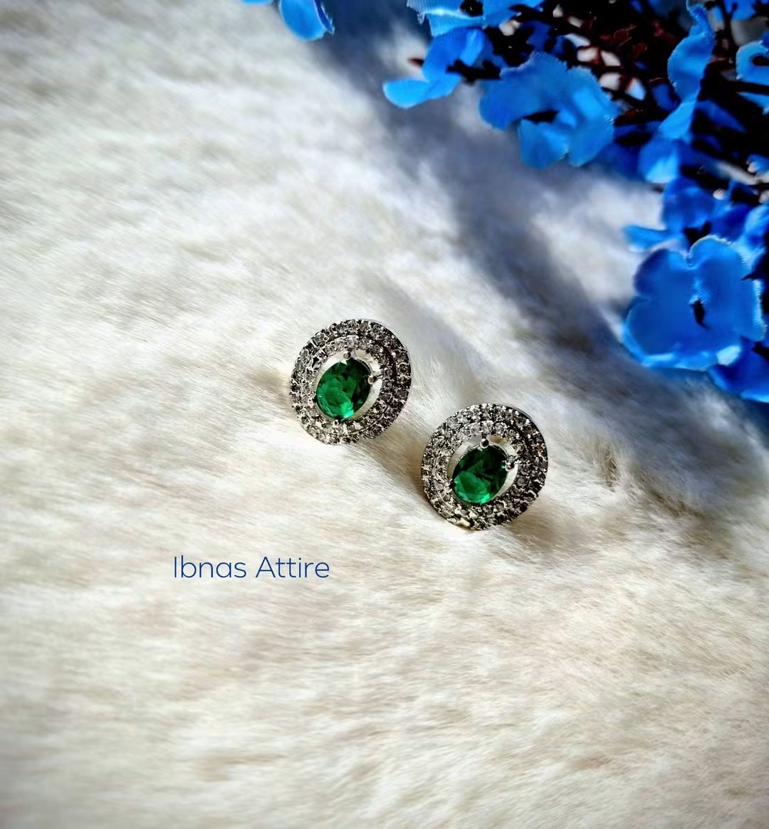 Silver-tone stud earrings featuring an oval emerald green center stone surrounded by a sparkling American diamond halo, laid flat on a textured white surface next to blue floral decor.