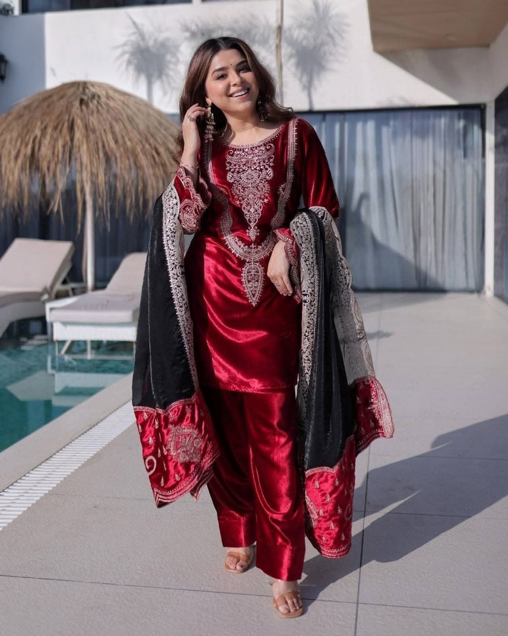 Woman wearing a luxurious maroon ice velvet long top and matching pants, styled with a contrasting dark decorative dupatta featuring ornate embroidery and a resort/poolside background.  