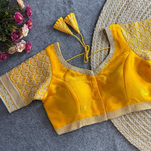 A yellow traditional blouse with ornate gold and silver scalloped lace sleeves and border trim, laid flat on a gray felt surface next to pink roses and a woven mat.