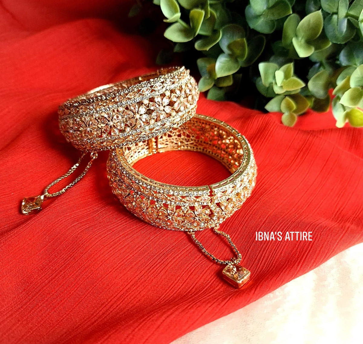 Pair of wide gold-plated bangles fully embellished with clear sparkling cubic zirconia stones and hanging stone dangles, resting on a bright red textured fabric next to green foliage.gold chains featuring sparkling cushion-cut stone drops attached to the openable screw pin.Smooth side hinge and screw mechanism that ensures easy wear without friction or squeezing hands.