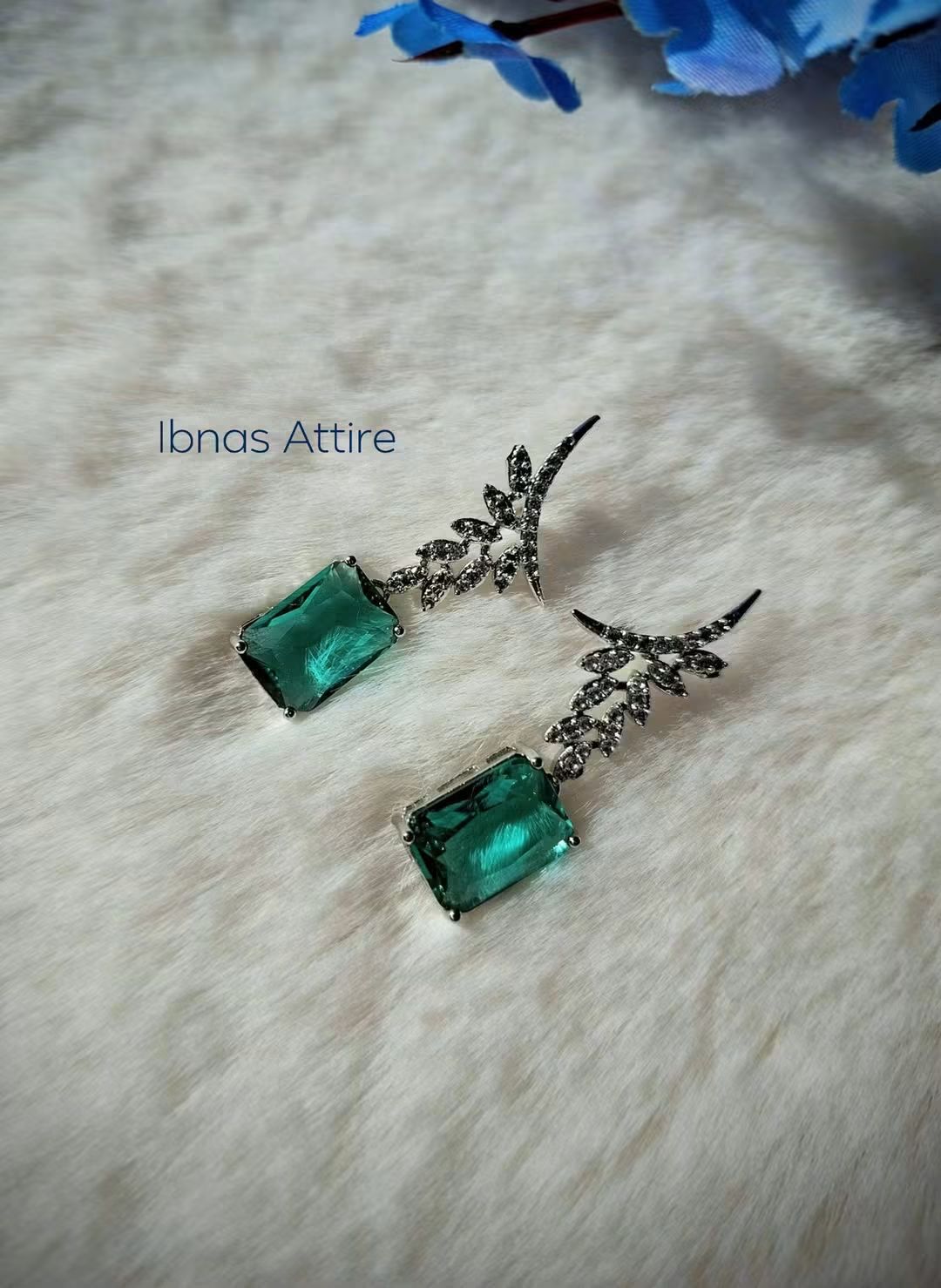 Silver-tone drop earrings with diamond-encrusted leaf posts and rectangular emerald green stone drops, displayed on a soft white fabric background with blue flower decor.
