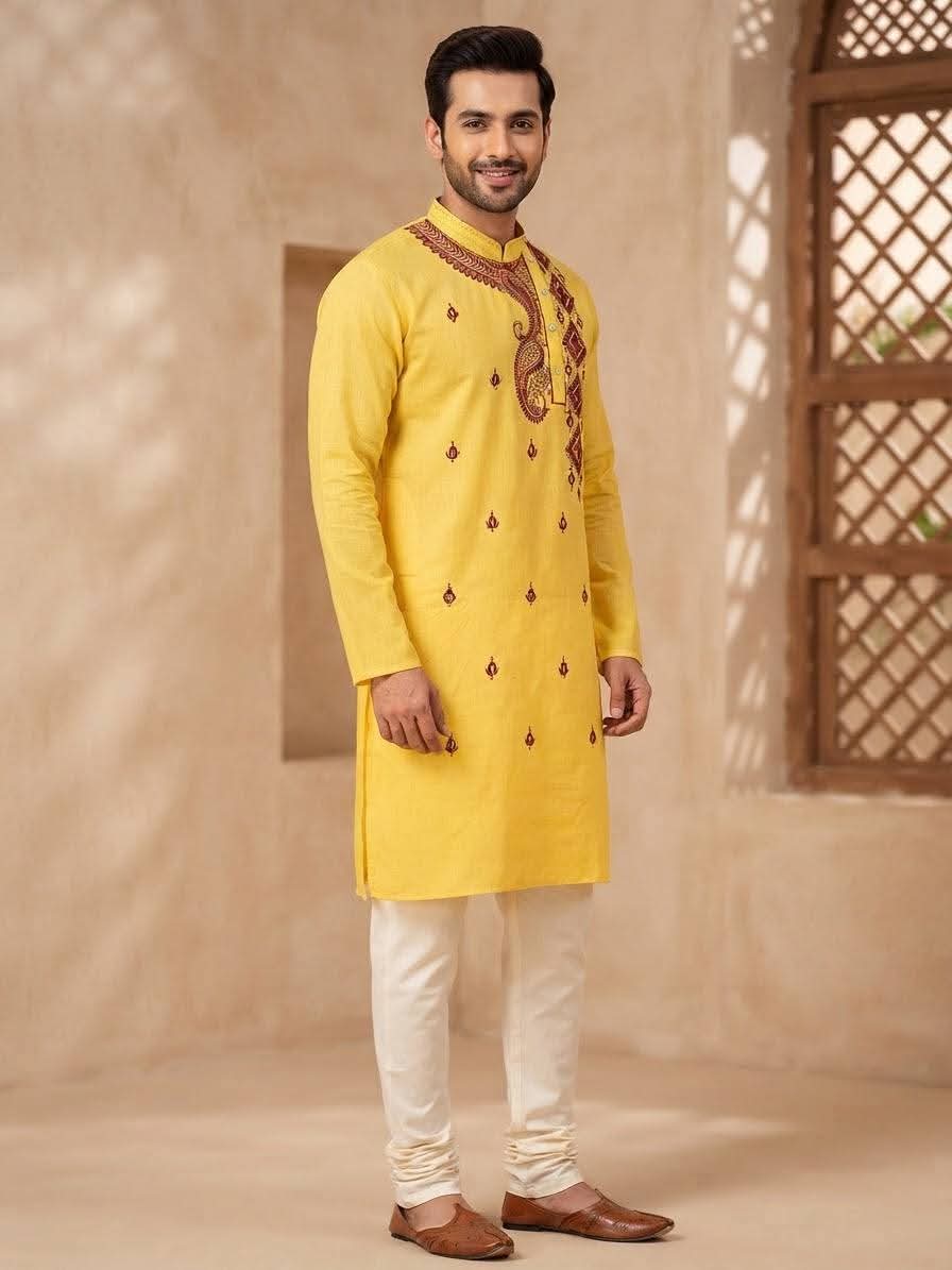 Model wearing a lemon yellow cotton long kurta with red/maroon embroidery, off-white churidar pants, brown leather shoes, rustic textured backdrop.