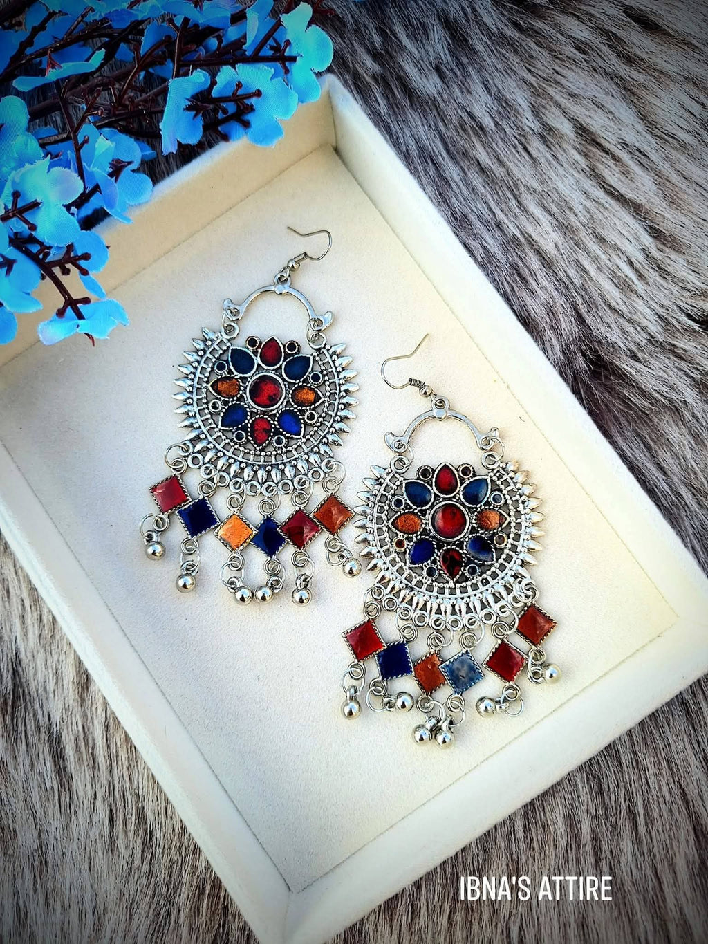 A pair of ornate silver-toned dangle earrings with colorful enamel details and dangling silver bells, displayed on a cream-colored jewelry box. The box is set on a gray faux fur surface with blue artificial flowers in the corner.