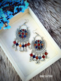 A pair of ornate silver-toned dangle earrings with colorful enamel details and dangling silver bells, displayed on a cream-colored jewelry box. The box is set on a gray faux fur surface with blue artificial flowers in the corner.
