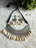 German silver rigid plate necklace with dangling cowrie shells, matching earrings with shell drops, black cord, laid flat on a textured white surface next to green foliage.