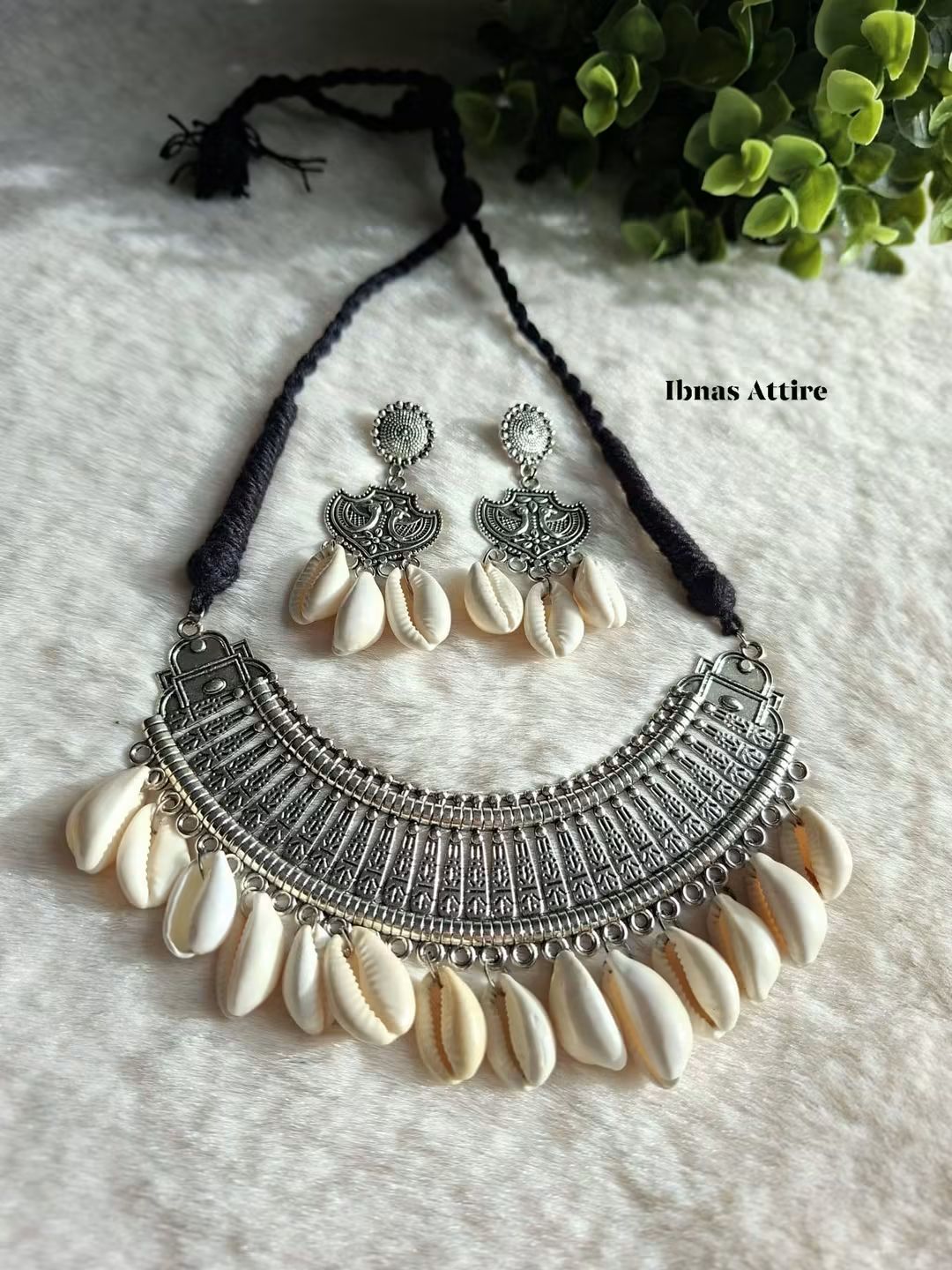 German silver rigid plate necklace with dangling cowrie shells, matching earrings with shell drops, black cord, laid flat on a textured white surface next to green foliage.
