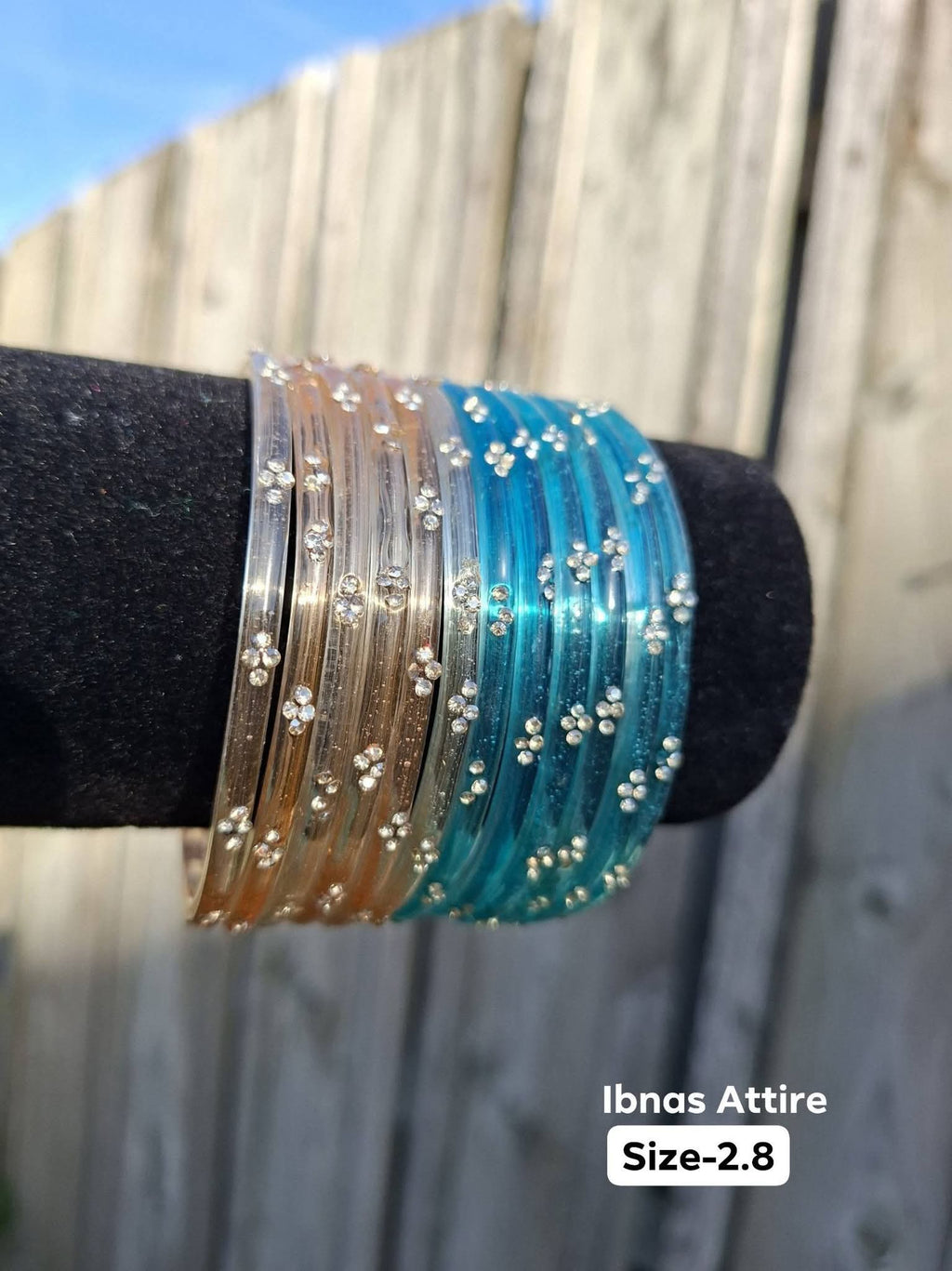 Stack of translucent glass bangles split between champagne gold and aqua cyan blue with gold sequin accents displayed on a black velvet bar outdoors against a wooden fence.