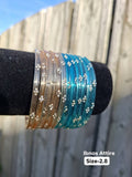 Stack of translucent glass bangles split between champagne gold and aqua cyan blue with gold sequin accents displayed on a black velvet bar outdoors against a wooden fence.