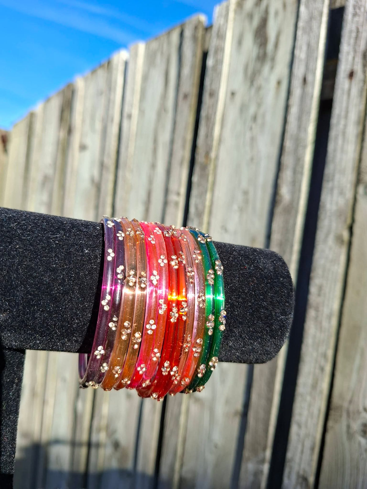 Stack of warm-toned multi-color glass bangles (purple, orange, red, pink, green) with gold sequin accents displayed on a black velvet bar outdoors against a wooden fence.