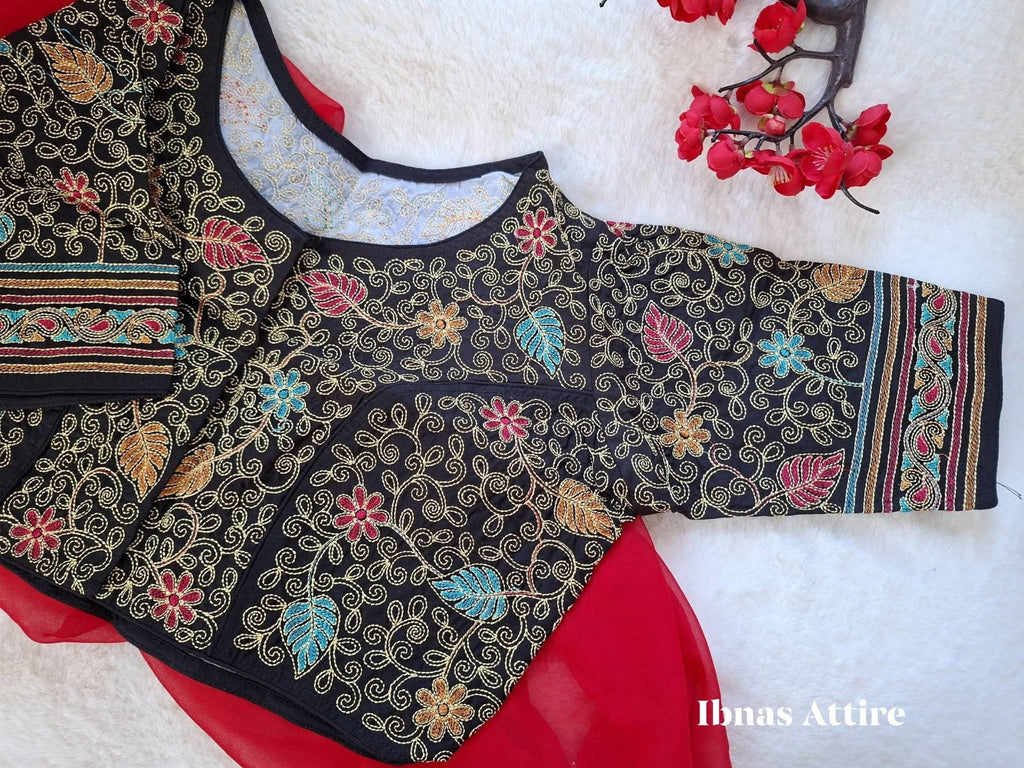 Black malai cotton blouse laid flat over a vibrant red drape on a soft white fur surface, displaying detailed multicolored Kantha embroidery and a front hook closure. Elegant geometric and paisley Kantha borders highlighting the sleeve cuffs and lower hem. 