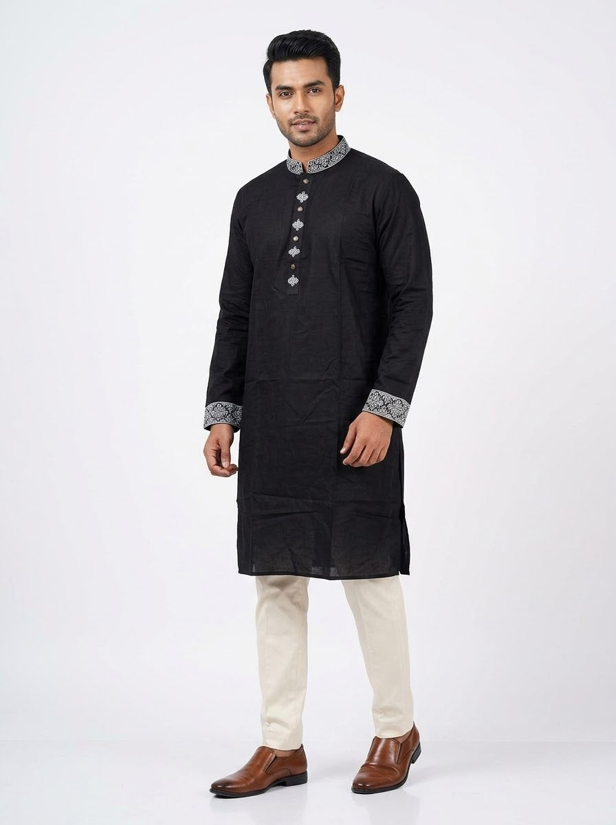 A man modeling a black long-sleeve kurta with silver patterned trim on the collar, cuffs, and front placket, paired with cream trousers and brown dress shoes, against a plain white studio background.