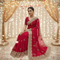 A woman seated indoors wearing a bright pink Designer Saree with Readymade Blouse. Vibrant pink/red crunchy texture saree,Statement scalloped/cutwork embroidery embedded with shimmering Swarovski crystals which Provides a unique tactile hand-feel and effortless volume control in pleats.