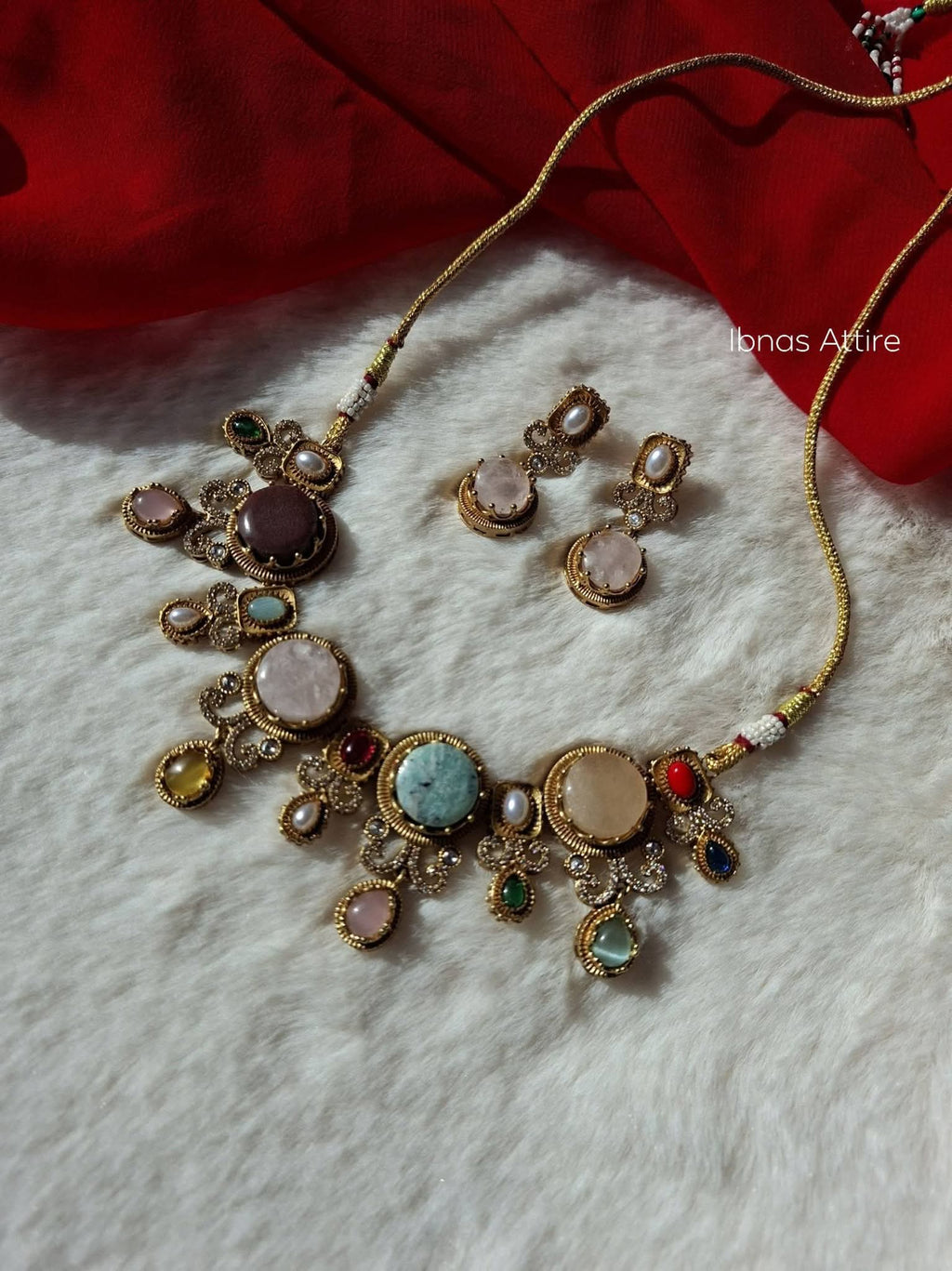 Antique gold-plated necklace set with oval and round multi-colored cabochon semi-precious stones and matching earrings displayed on a white faux-fur backdrop beside a red drape.Bezel-set round cabochon stones in complementary earth and pastel tones balanced across textured antique gold frames.Subtle pearl droplets and delicate cubic zirconia/crystal halo trims surrounding each central stone.  Soft woven gold cord allowing you to wear it as a close-fit choker or a longer princess-length necklace.
