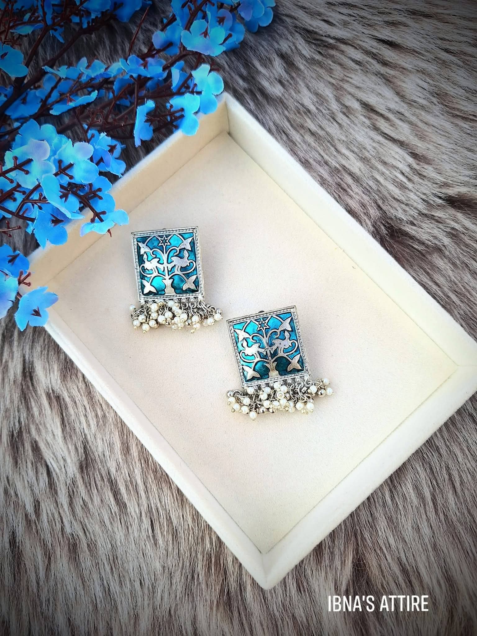 A pair of square silver-toned earrings with turquoise and white enamel detailing and dangling pearl-like beads, displayed on a cream tray over a gray faux-fur surface with blue flowers in the corner. 