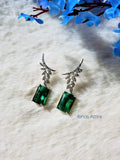 Silver-tone drop earrings with diamond-encrusted leaf posts and rectangular emerald green stone drops, displayed on a soft white fabric background with blue flower decor.