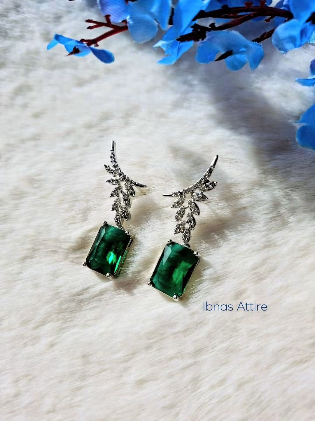 Silver-tone drop earrings with diamond-encrusted leaf posts and rectangular emerald green stone drops, displayed on a soft white fabric background with blue flower decor.