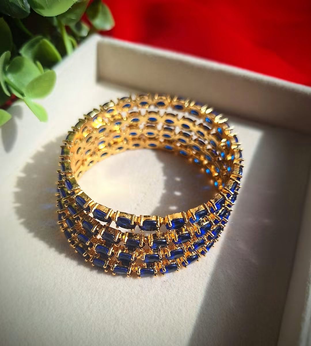 A gold-plated 4 piece of American Diamond Blue bangle bracelet decorated with rectangular blue gemstones, displayed on a light beige jewelry box with a red background and green leaves in the corner.