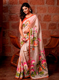 A woman wearing a beige linen digital printed  saree with vibrant pink, orange, and green lotus floral prints, standing in front of a rustic brick wall and wooden chairs.