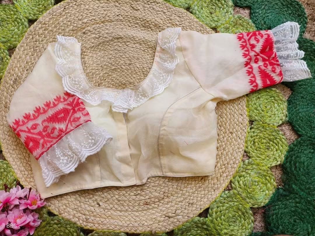 A cream-colored resham jamdani Readymade blouse with white lace ruffles and red embroidered sleeve cuffs, laid flat on a round woven straw mat over a green textured rug.