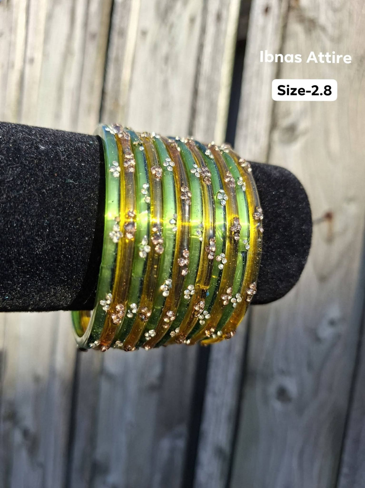 Stack of translucent olive green and golden amber glass bangles with gold sequin accents displayed on a black velvet bar outdoors against a wooden fence. Contrast between fresh olive green and warm amber glass rings.