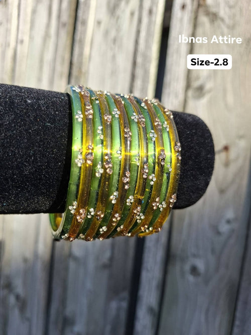 Stack of translucent olive green and golden amber glass bangles with gold sequin accents displayed on a black velvet bar outdoors against a wooden fence. Contrast between fresh olive green and warm amber glass rings.