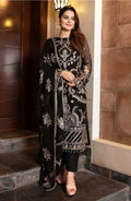Woman wearing a black luxury embroidered long Pakistani tunic with matching dupatta and trousers, posed against a dark doorway background.Rich black chiffon base highlighted by fine silver-toned traditional zari and glittering sequins work.