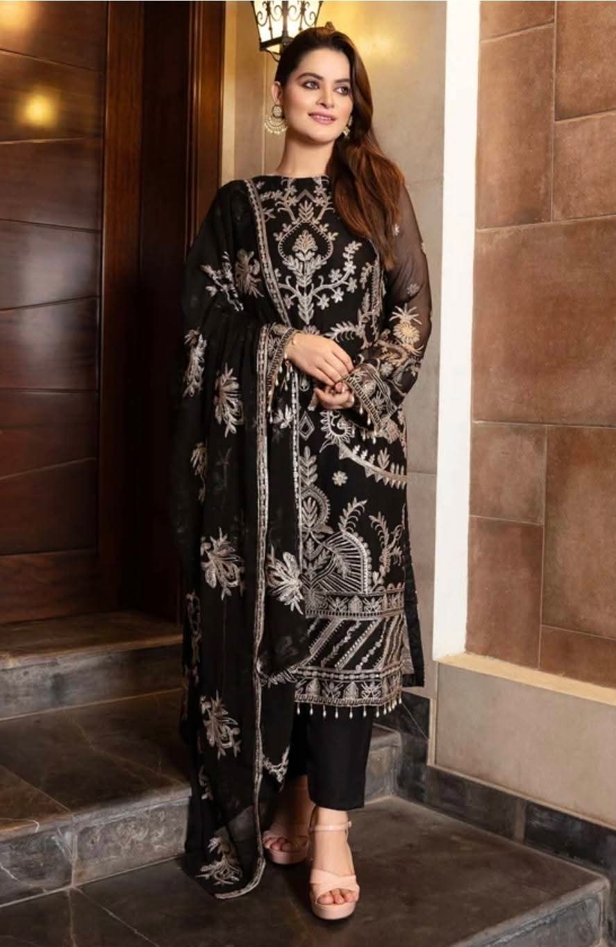 Woman wearing a black luxury embroidered long Pakistani tunic with matching dupatta and trousers, posed against a dark doorway background.Rich black chiffon base highlighted by fine silver-toned traditional zari and glittering sequins work.