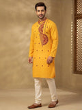 Model wearing a marrigold color cotton long kurta with red/maroon embroidery, off-white churidar pants, brown leather shoes, rustic textured backdrop.