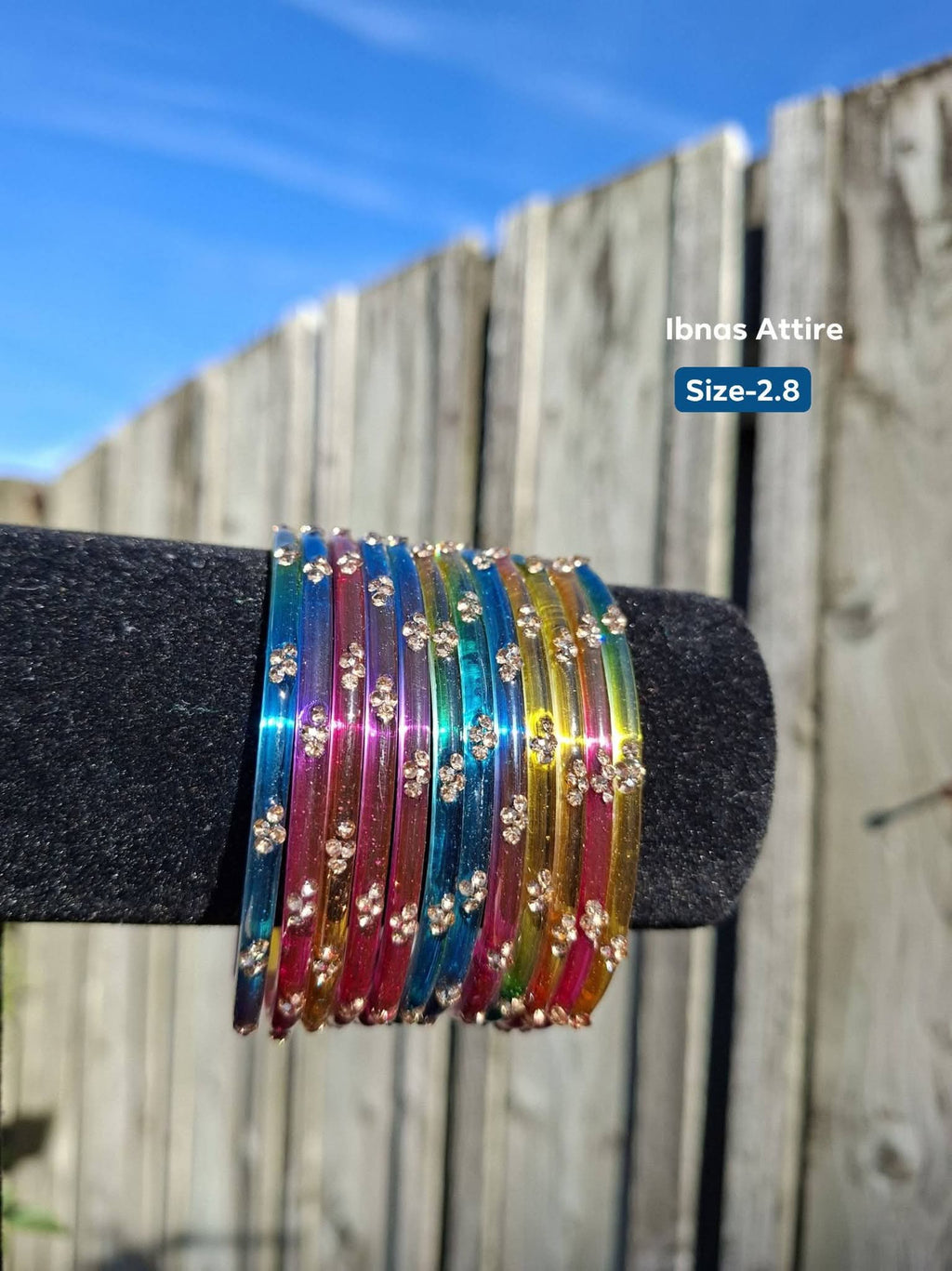 Stack of vibrant multi-colored glass bangles (blue, pink, green, yellow) with gold sequin accents displayed outdoors on a black velvet bracelet holder. 
