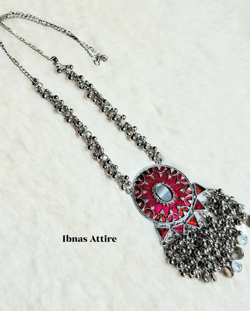 Oxidized silver long necklace laid on a white textured background, featuring a round pendant with a mirror and red enamel details, accented with metallic bead fringes.Intricately detailed silver bead chains provide a layered, traditional look.