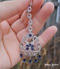 Close-up of a silver American diamond maang tikka featuring clear pave-set stones and royal blue teardrop crystal drops held against an outdoor background.Striking blue stone center flower and matching dangling drops for a regal pop of color. 