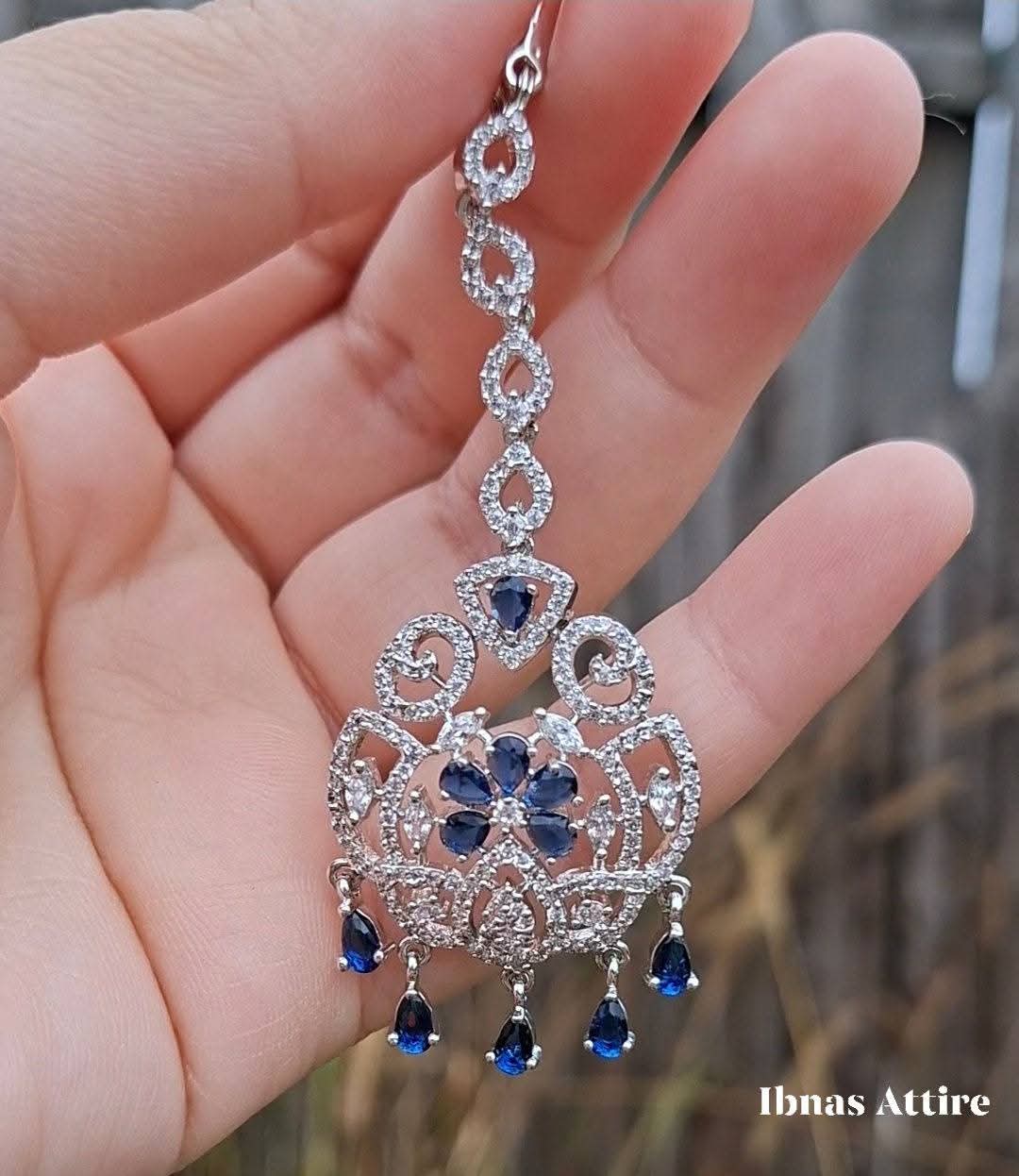 Close-up of a silver American diamond maang tikka featuring clear pave-set stones and royal blue teardrop crystal drops held against an outdoor background.Striking blue stone center flower and matching dangling drops for a regal pop of color. 