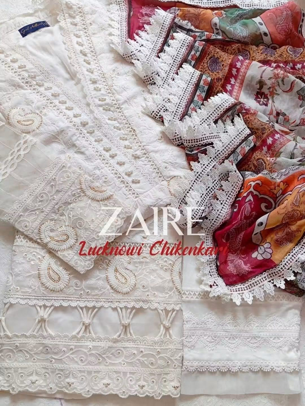 A folded white Stitched Luxury Chikankari 3-Piece Suit-Fine white-on-white thread embroidery complemented by intricate patterns and pearl embellishments.A vibrant multi-color contrasting print framed elegantly with a 4-sided detailed lace stitch.Clean, matching tailored trousers designed for effortless comfort and balance.
