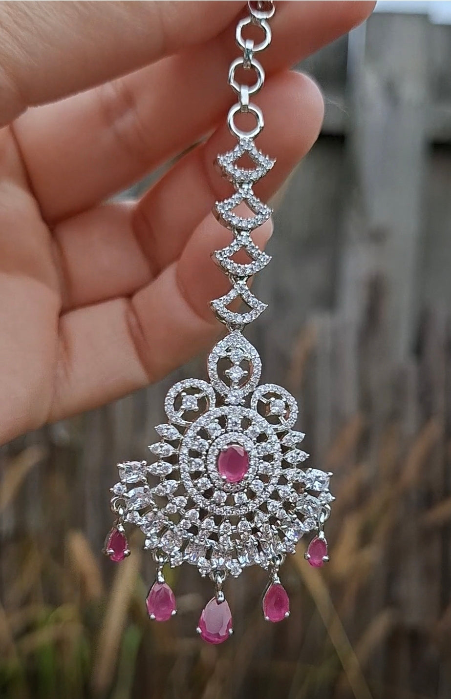 Close-up of a hand holding a silver American diamond maang tikka featuring clear pave stones and ruby red drop crystals against an outdoor backdrop. High-clarity stones offering maximum brilliance and sparkle.