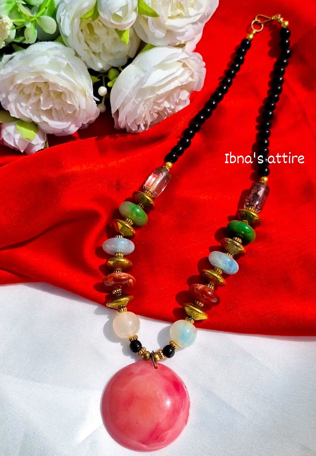 Featuring a Boho-Chic / Artisan Statement Necklace-Sleek upper chain made of small dark beads that sits comfortably on the neck.A smooth, oversized circular pink stone centerpiece that anchors the design with a vibrant splash of color.Asymmetrical arrangement of green, blue, reddish-brown, and translucent beads separated by rustic gold-tone spacer coins.