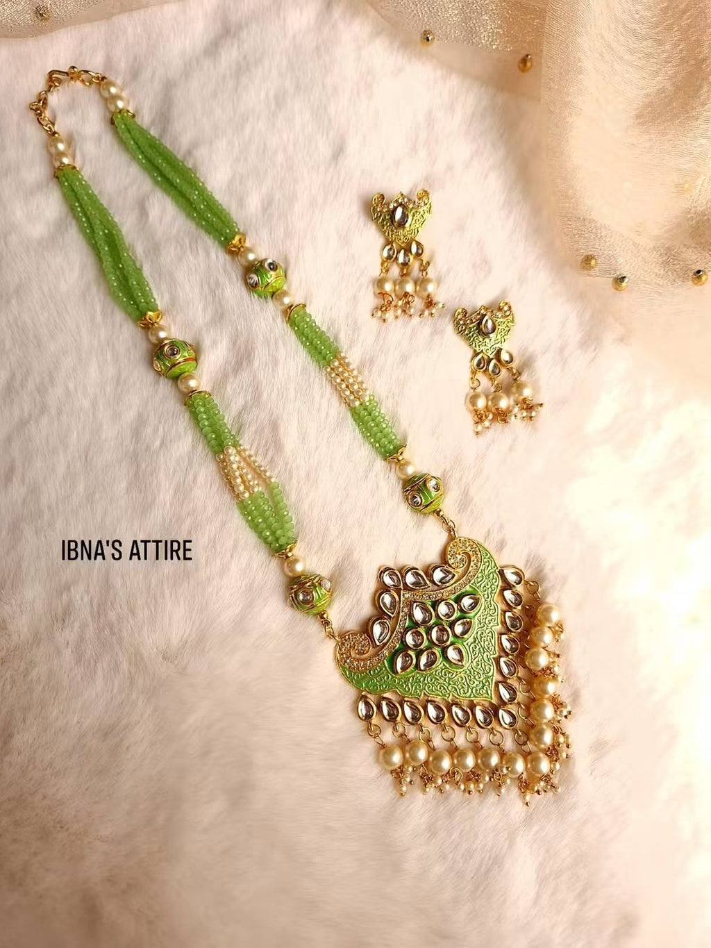 A green and gold traditional Indian kundan jewelry set featuring a necklace and matching earrings, displayed on a soft white faux fur surface. The necklace has a green beaded chain with gold-toned embellishments and a large gold-toned pendant decorated with green stones and pearl-like beads.